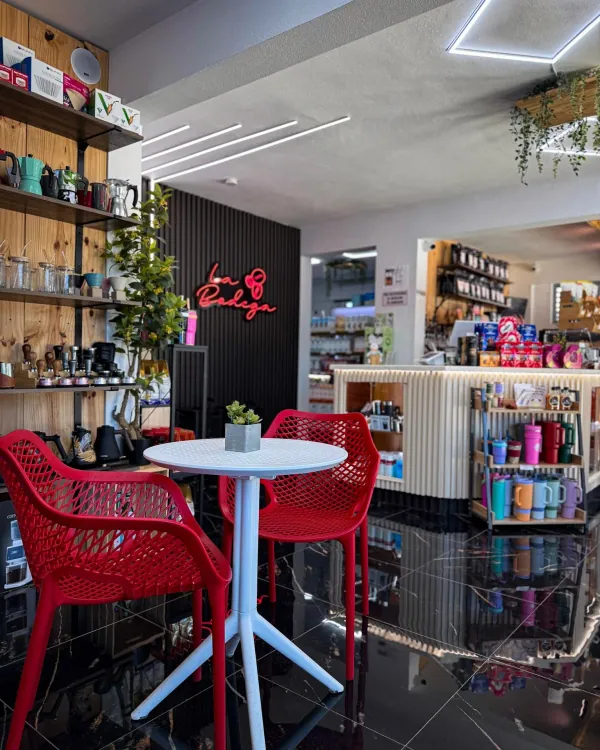 Interior de La Bodega: mesa blanca con dos sillas rojas, estantes de madera con cafeteras y el letrero de neón de La Bodega